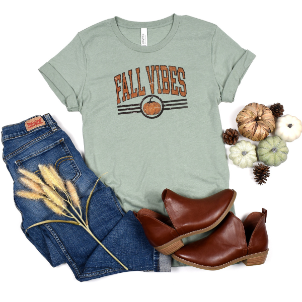 A green t-shirt with 'FALL VIBES' text and an autumn-themed design, paired with blue jeans, brown ankle boots, and decorative pine cones.