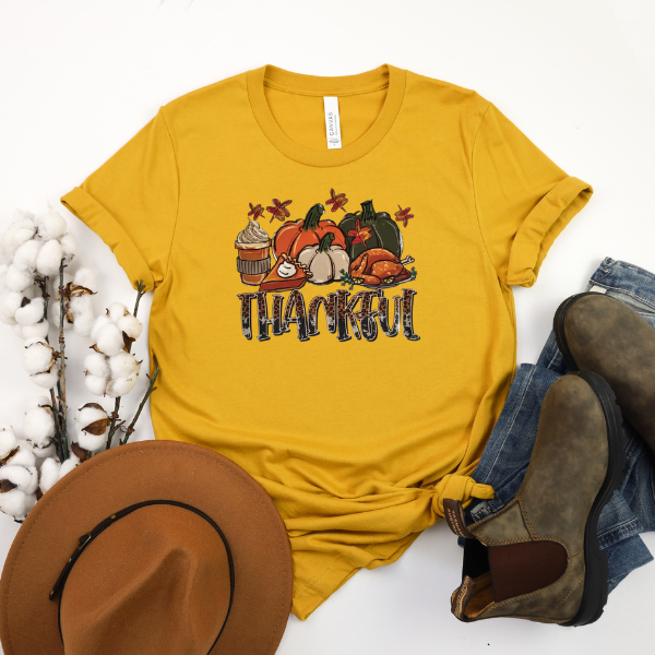 A yellow t-shirt with 'THANKFUL THANKSGIVING' text and a graphic of pumpkins, cranberry sauce, and stuffing, displayed flat with a pair of brown boots and a hat on a white background.