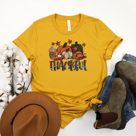 A yellow t-shirt with 'THANKFUL THANKSGIVING' text and a graphic of pumpkins, cranberry sauce, and stuffing, displayed flat with a pair of brown boots and a hat on a white background.