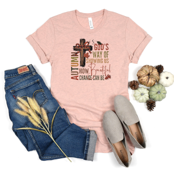A pink shirt with text 'Autumn is God's way of showing us how beautiful change can be' and a cross symbol, paired with blue jeans and a pair of grey shoes, accompanied by decorative pine cones and a dry plant stem.