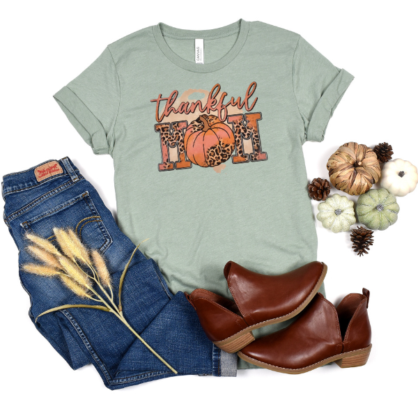 A green t-shirt with 'Thankful' and a pumpkin design, paired with blue jeans and brown ankle boots, accompanied by pine cones and dry plants.