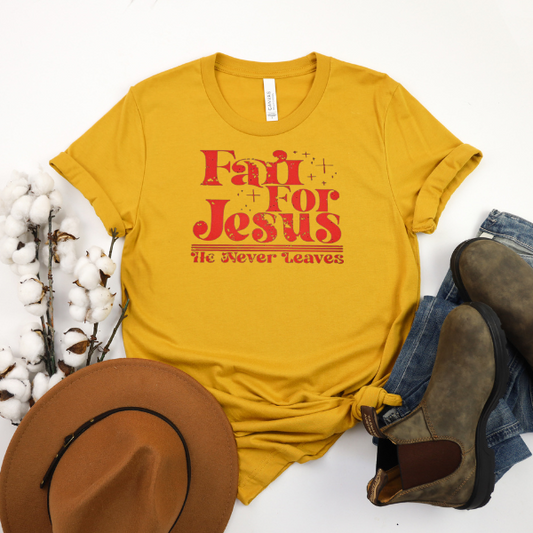 A yellow t-shirt with the text 'FAIL FOR JESUS THE NEVER LEAVES' printed on the front, displayed flat with a pair of brown boots and a hat on a white surface, accompanied by cotton bolls and decorative tobacco leaves.