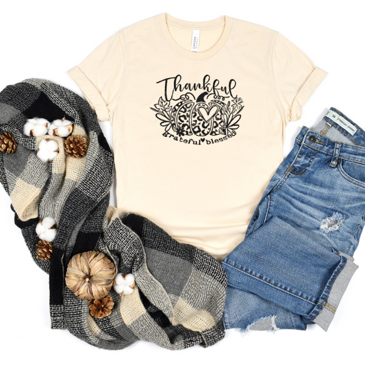 A beige t-shirt with 'Thankful, Grateful, Blessed' printed on the front, accompanied by a blue scarf, white cotton pants, and a pine cone decoration.