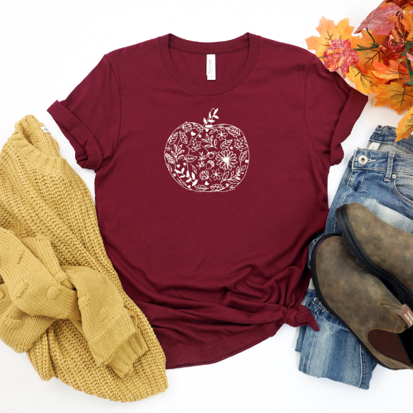 A maroon colored shirt with a decorative pumpkin design, paired with blue jeans and brown boots, accompanied by a knit hat and leaf accents.