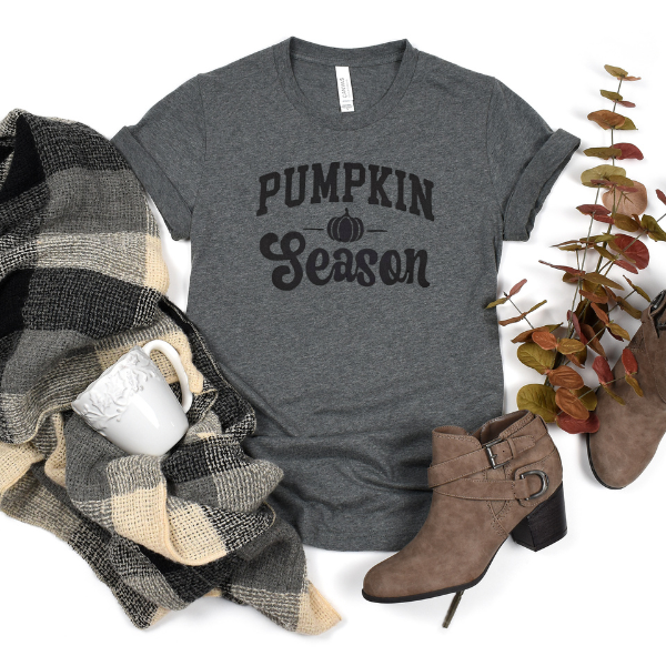 A gray t-shirt with the text 'PUMPKIN SEASON' printed on the front, accompanied by a plaid scarf, a pair of brown boots, and decorative leaves.