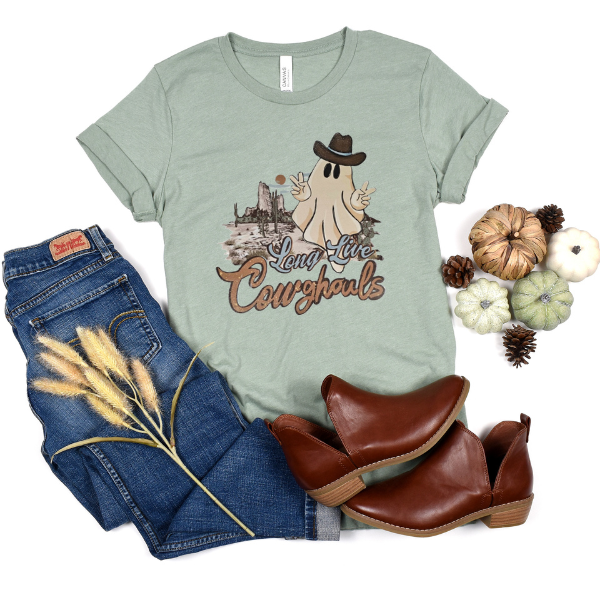 A green t-shirt with 'Long Live Cowghouls' text and a graphic of a ghost wearing a cowboy hat, accompanied by blue jeans, brown shoes, and decorative pine cones.